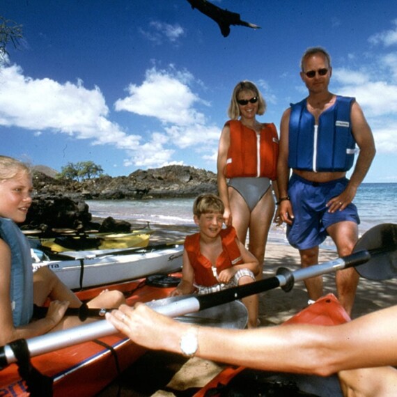 A family at the beach, smiling and preparing to kayak, with ocean and sand in the background.