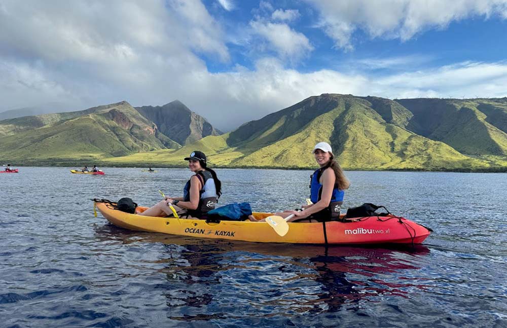 Two people in a kayak on the ocean, with impressive mountains providing a picturesque backdrop.