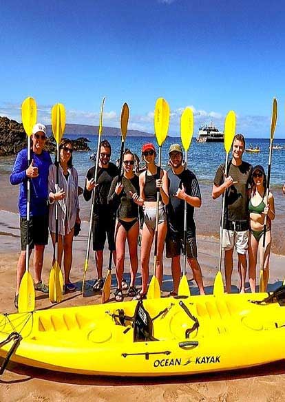 A Group of individuals stands on the west Maui beach, smiling and posing with colorful kayaks beside them.