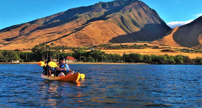 Kayakers wearing life jackets paddling on calm ocean waters in Maui while following Ocean safety guidelines Hawaii near a mountainous coastline.