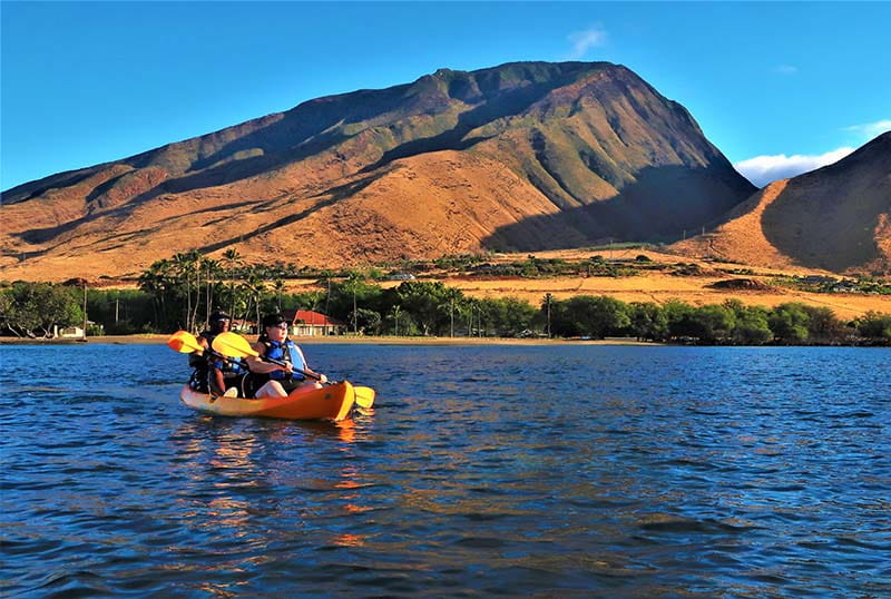 Kayakers wearing life jackets paddling on calm ocean waters in Maui while following Ocean safety guidelines Hawaii near a mountainous coastline.